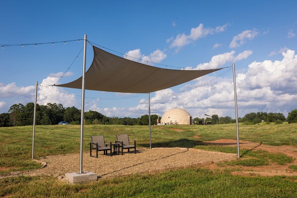 A tent-like structure with a table and chairs, a dome-shaped building in the background, and a grassy field.