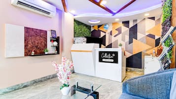 A reception area with a reception desk, a wall-mounted air conditioner, a glass coffee table, a blue sofa, and a decorative plant.