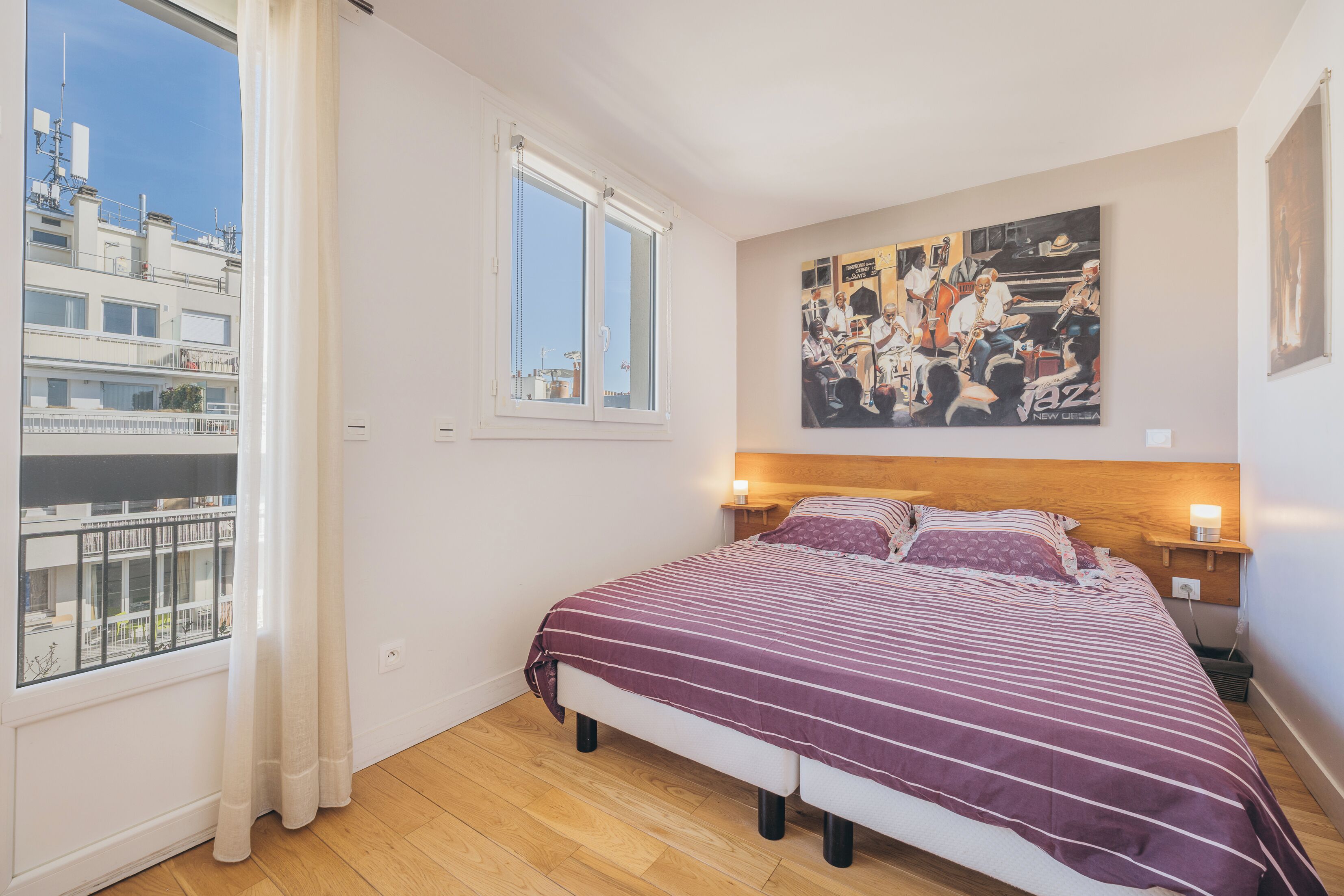 Une chambre avec un grand lit, une fenêtre avec des rideaux, un tableau au mur et une vue sur les bâtiments à l’extérieur.