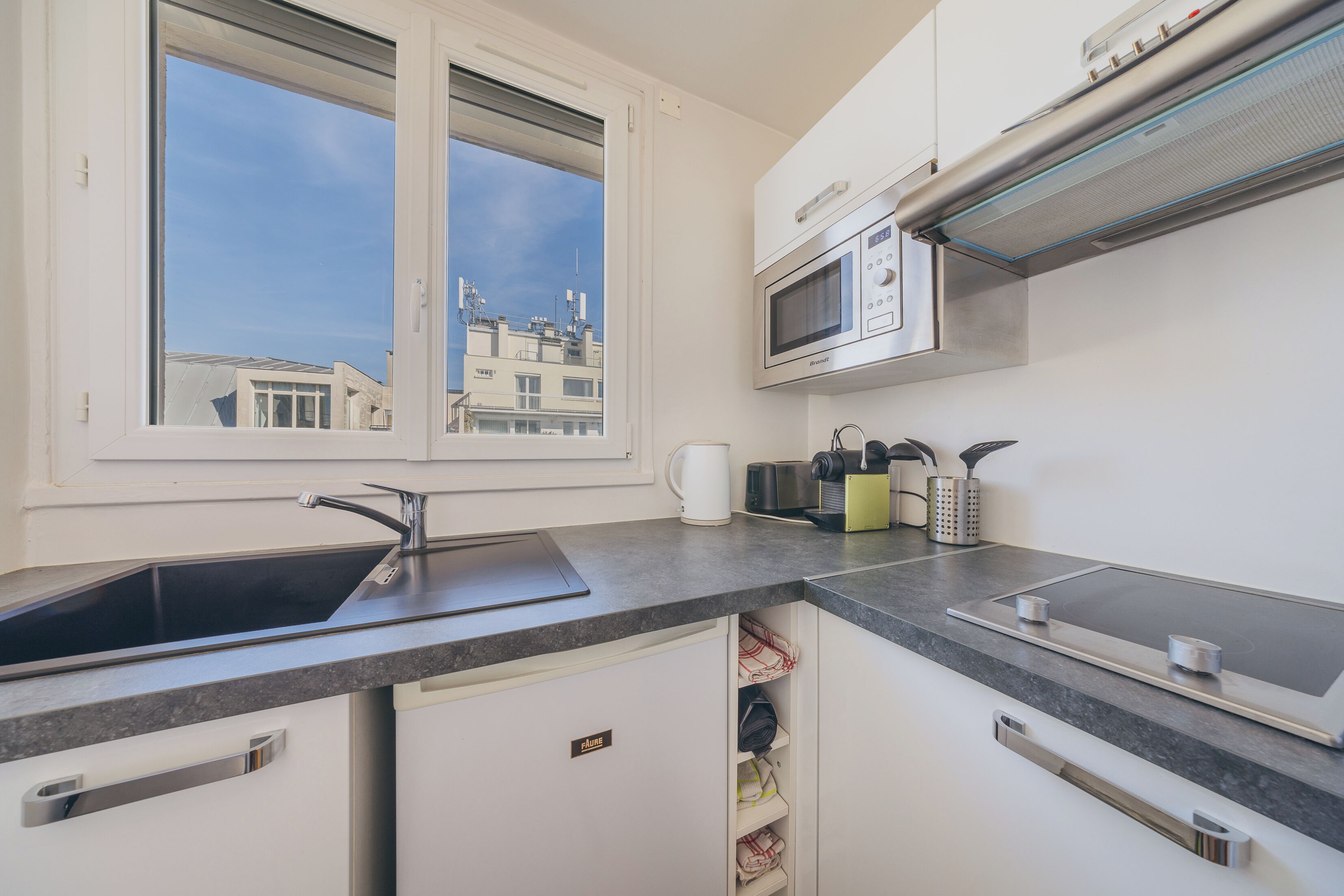 Une cuisine moderne équipée d’un four à micro-ondes encastré, d’un évier et d’une fenêtre donnant sur des immeubles.