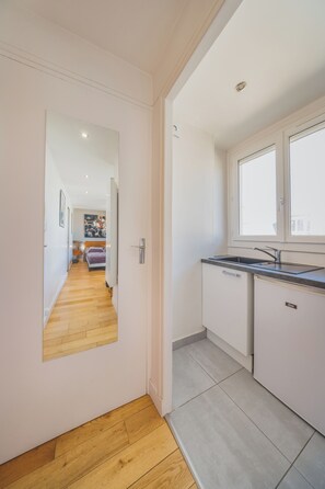 A narrow hallway leading to a bedroom with a large mirror, a wooden floor, and a kitchen with a window and sink.