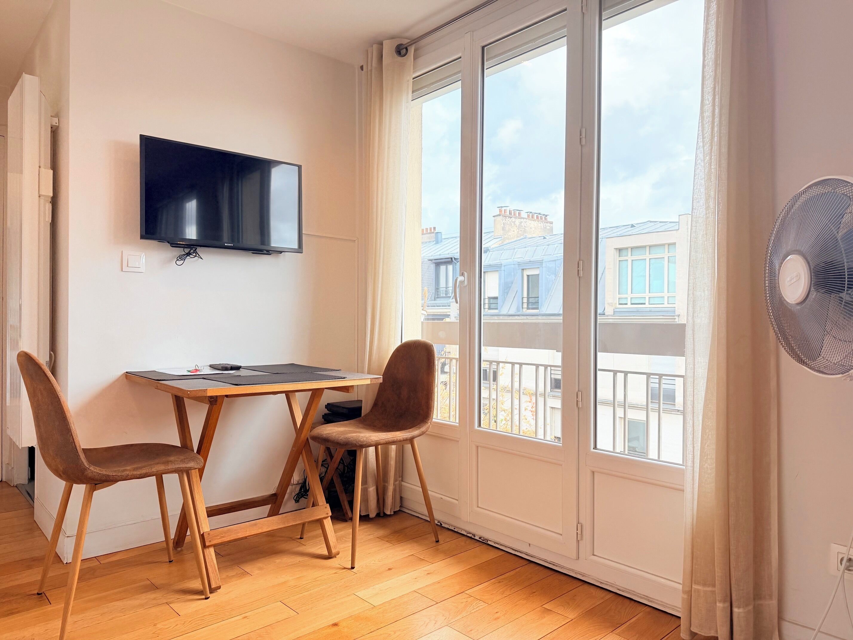 Une pièce avec une table en bois, deux chaises, un téléviseur à écran plat fixé au mur et une grande fenêtre donnant sur des immeubles.