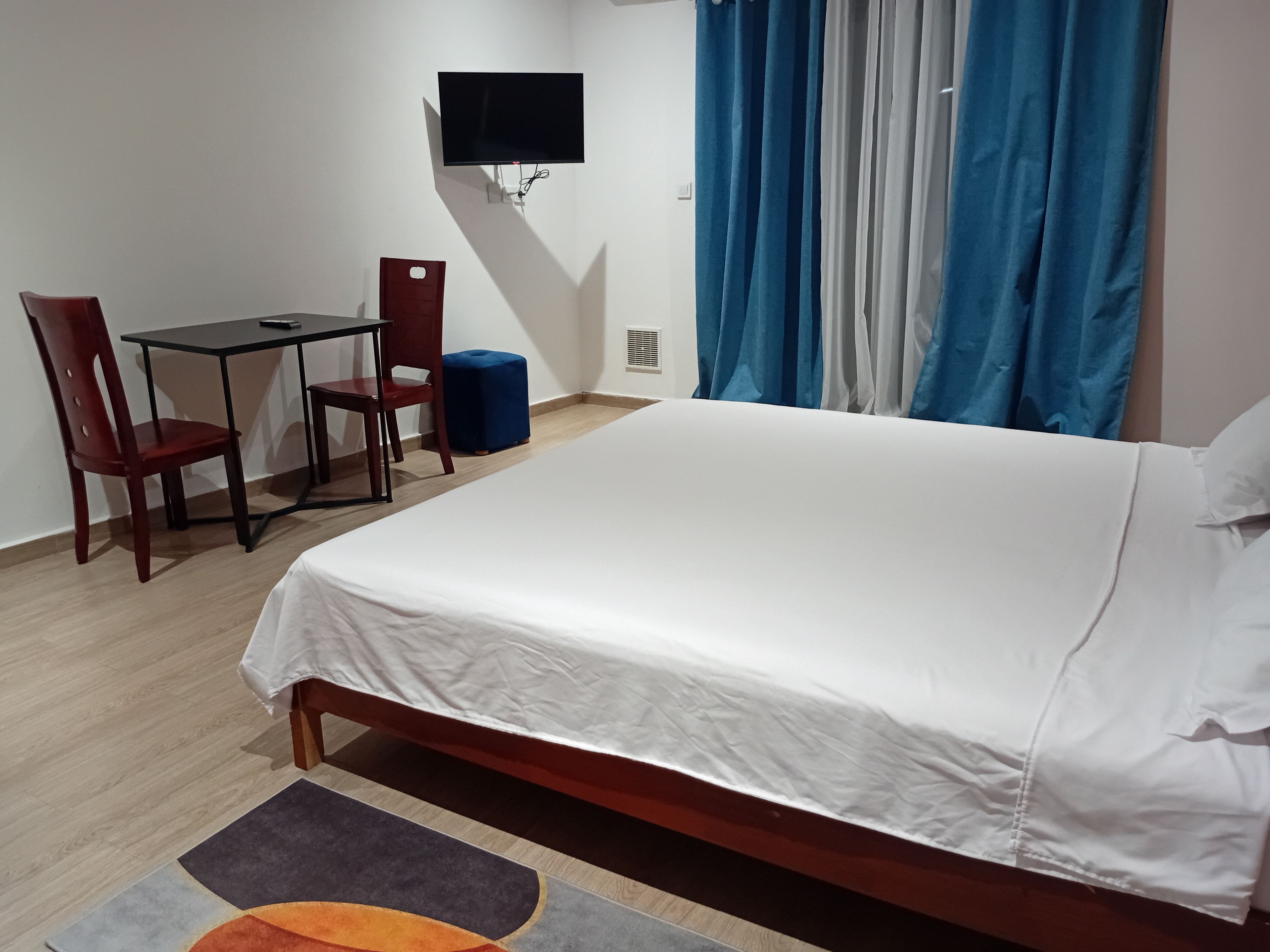 Une chambre d’hôtel avec un lit, un bureau, deux chaises, un téléviseur fixé au mur et une fenêtre avec des rideaux bleus.