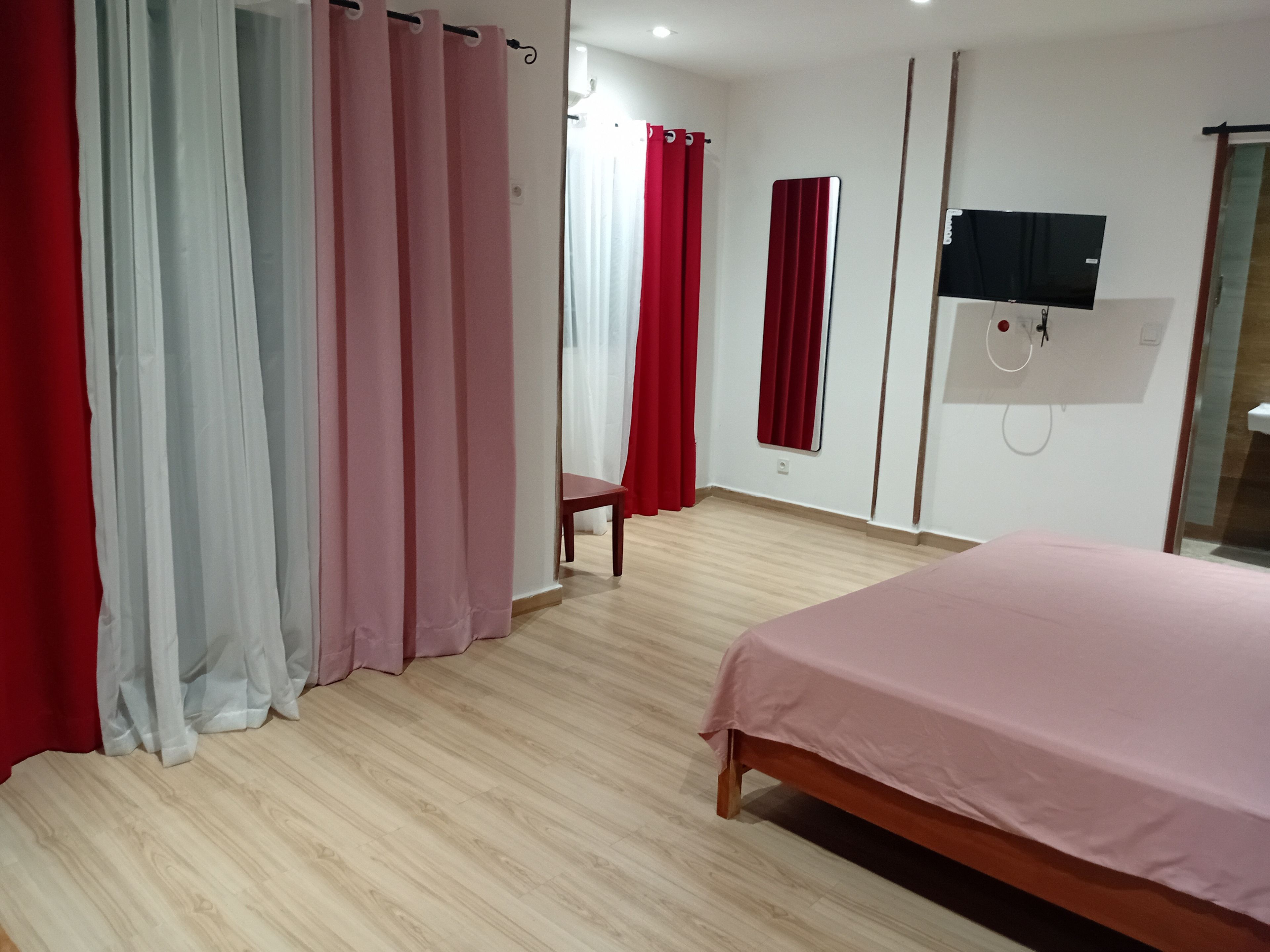Een slaapkamer met een bed, een houten vloer, een aan de muur bevestigde televisie en rode gordijnen.