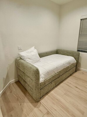 A small, single-bed room with a grey upholstered bed, a white pillow, and a light-colored wooden floor. - Spacious Peoria retreat with private pool, 5 bedrooms & family fun! (Peoria)