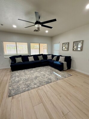 A living room with a sectional sofa, a ceiling fan, and a large window with blinds. - Spacious Peoria retreat with private pool, 5 bedrooms & family fun! (Peoria)