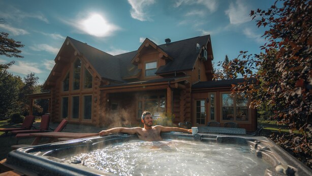 A person relaxing in a hot tub in front of a large log cabin with a chimney.