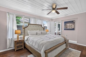 A bedroom with a wooden bed, two bedside lamps, a ceiling fan, and a large window with curtains. - Kylia Kove Family Retreat Minutes From Anna Maria Island (Bradenton)