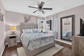 A bedroom with a bed, a ceiling fan, a nightstand with a lamp, a window with curtains, and a mirror.