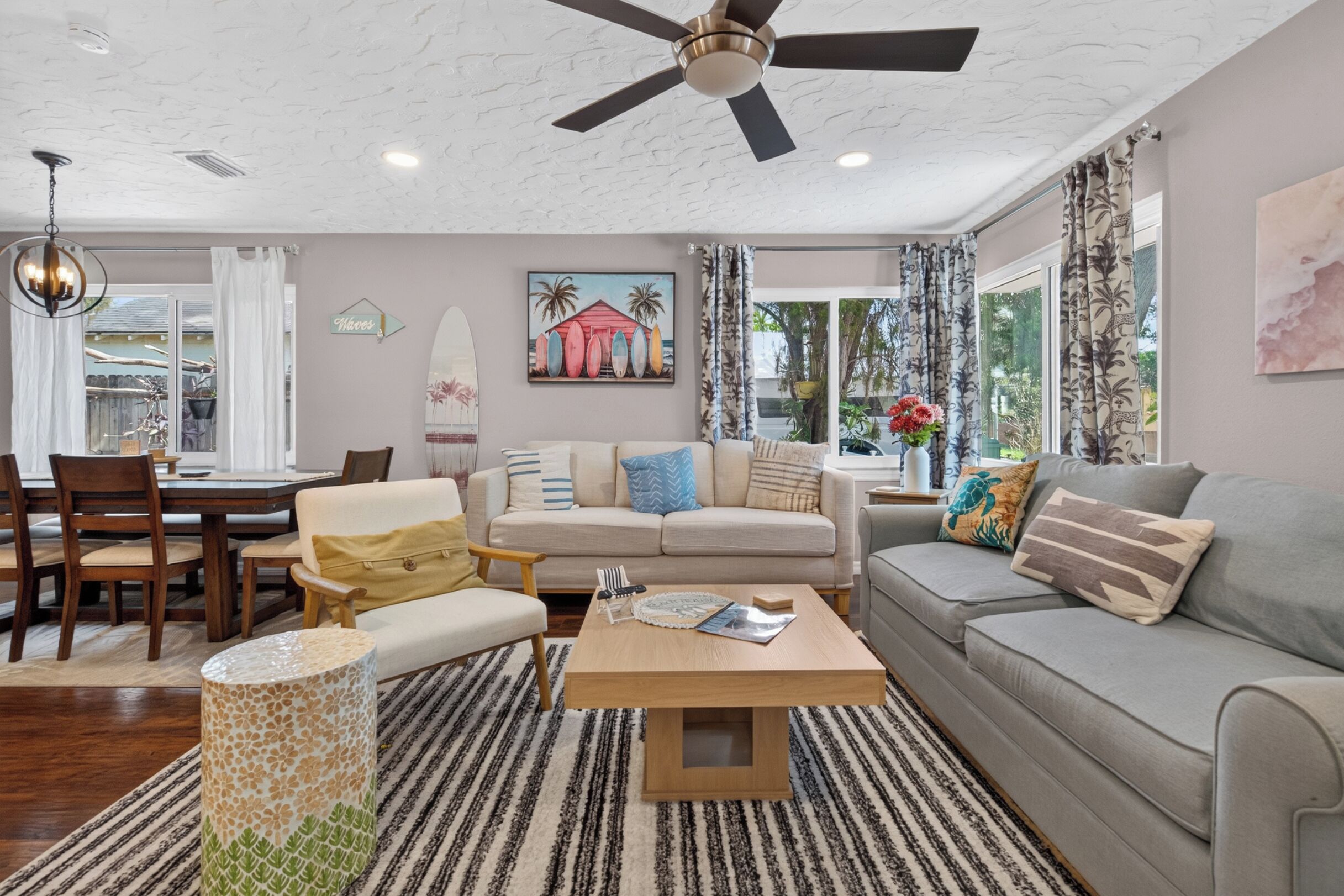 A living room with a ceiling fan, a dining area with a table and chairs, and a view of the outdoors through large windows.