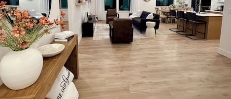 A spacious living area with a wooden floor, a large mirror, a sofa, a coffee table, and a dining area with chairs and a table.