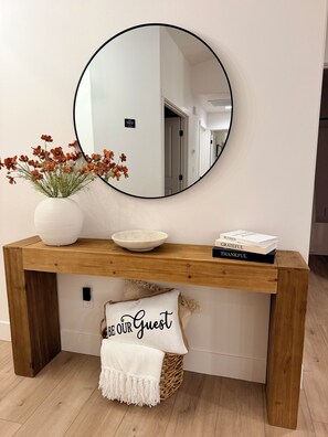 Une console en bois surmontée d’un miroir rond, avec un vase de fleurs, un bol et une pile de livres.