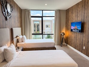 A hotel room with two beds, a TV on the wall, and a large window with a view of buildings.