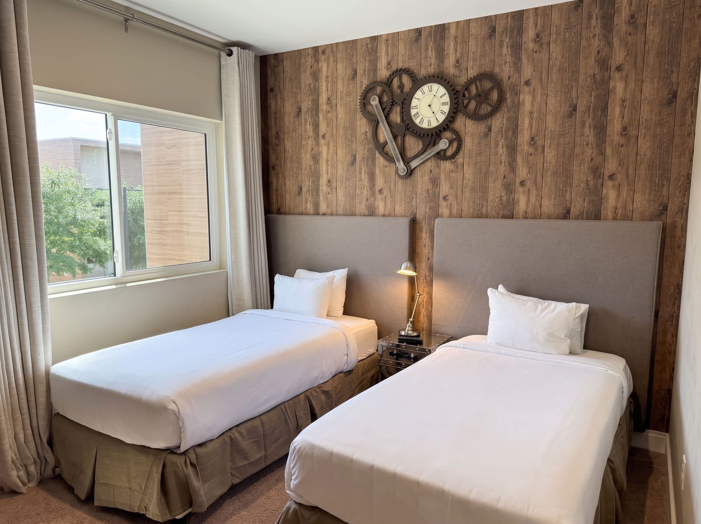 Une chambre d’hôtel avec deux lits, un mur en bois orné d’une horloge et une fenêtre avec des rideaux.