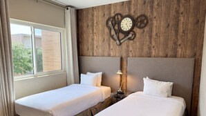 A hotel room with two beds, a wooden wall with a clock, and a window with curtains. - Magic Village Paradise 4BR (Kissimmee)