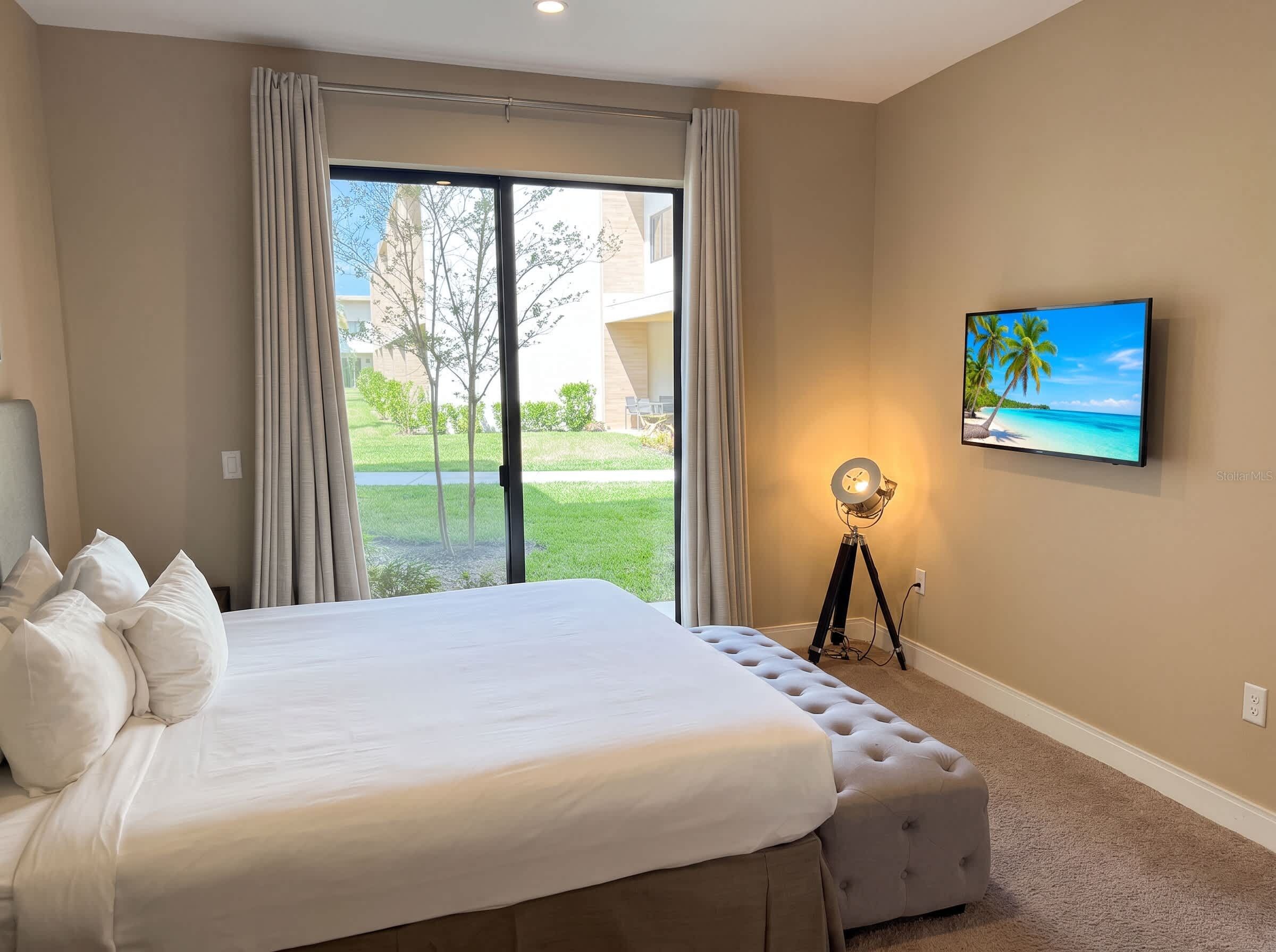 Une chambre d’hôtel avec un grand lit, un téléviseur à écran plat fixé au mur et une vue sur un immeuble et la verdure environnante.