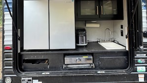 A compact RV kitchen with a refrigerator, stove, sink, and a GreyStone entertainment system.