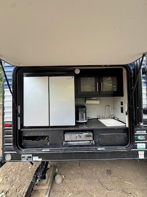 A compact RV kitchen with a refrigerator, stove, sink, and a GreyStone entertainment system.