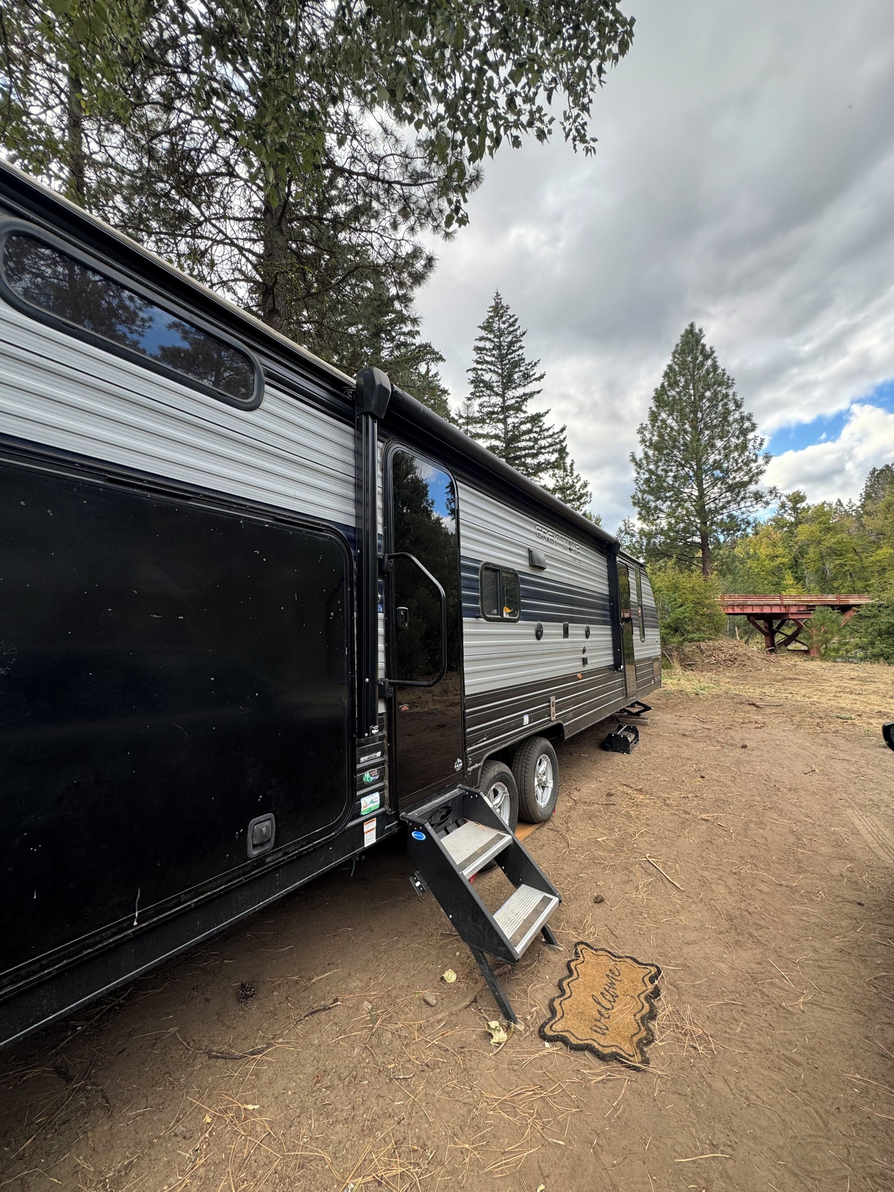 A black RV with a ramp and a welcome mat in a wooded area.