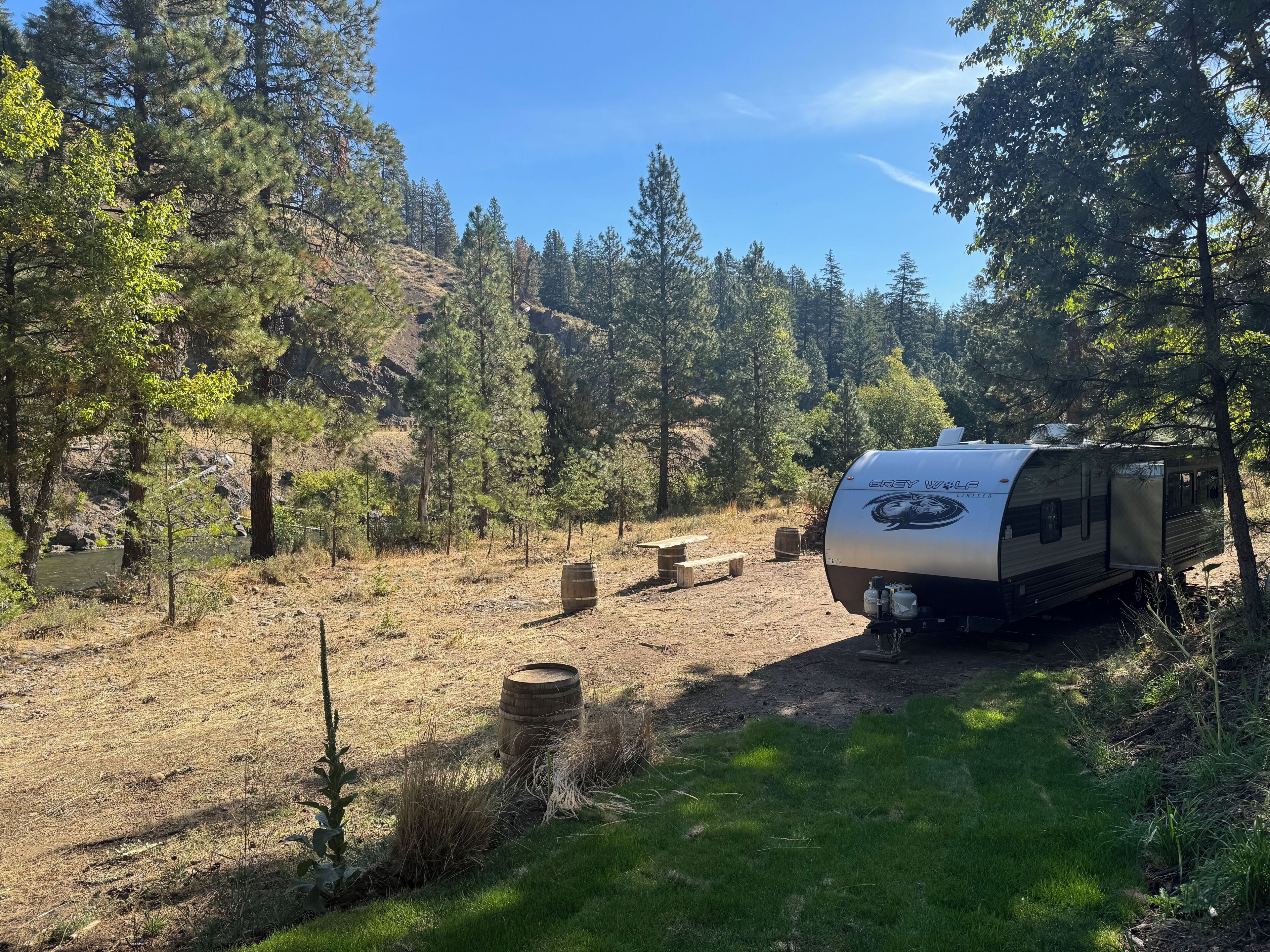 A Grey Wolf RV parked in a forested area with a picnic table and a barrel nearby.