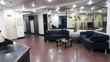 Un área de recepción moderna con un sofá negro, una mesa de centro y un mostrador de recepción. Hay plantas en macetas y un espejo grande en la pared.