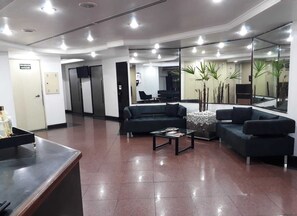 A modern reception area with a black sofa, a coffee table, and a reception desk. There are potted plants and a large mirror on the wall. - Apartamento GoodLar Liberdade SP  (Liberdade São Paulo)