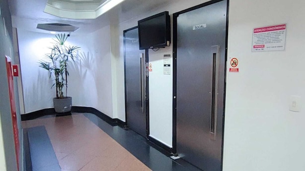 A corridor with a potted plant, a flat-screen TV mounted on the wall, and a door labeled "SOCIAL". - Apartamento GoodLar Liberdade SP  (Liberdade São Paulo)