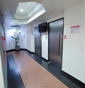 A corridor with a potted plant, a flat-screen TV mounted on the wall, and a door labeled "SOCIAL". - Apartamento GoodLar Liberdade SP  (Liberdade São Paulo)