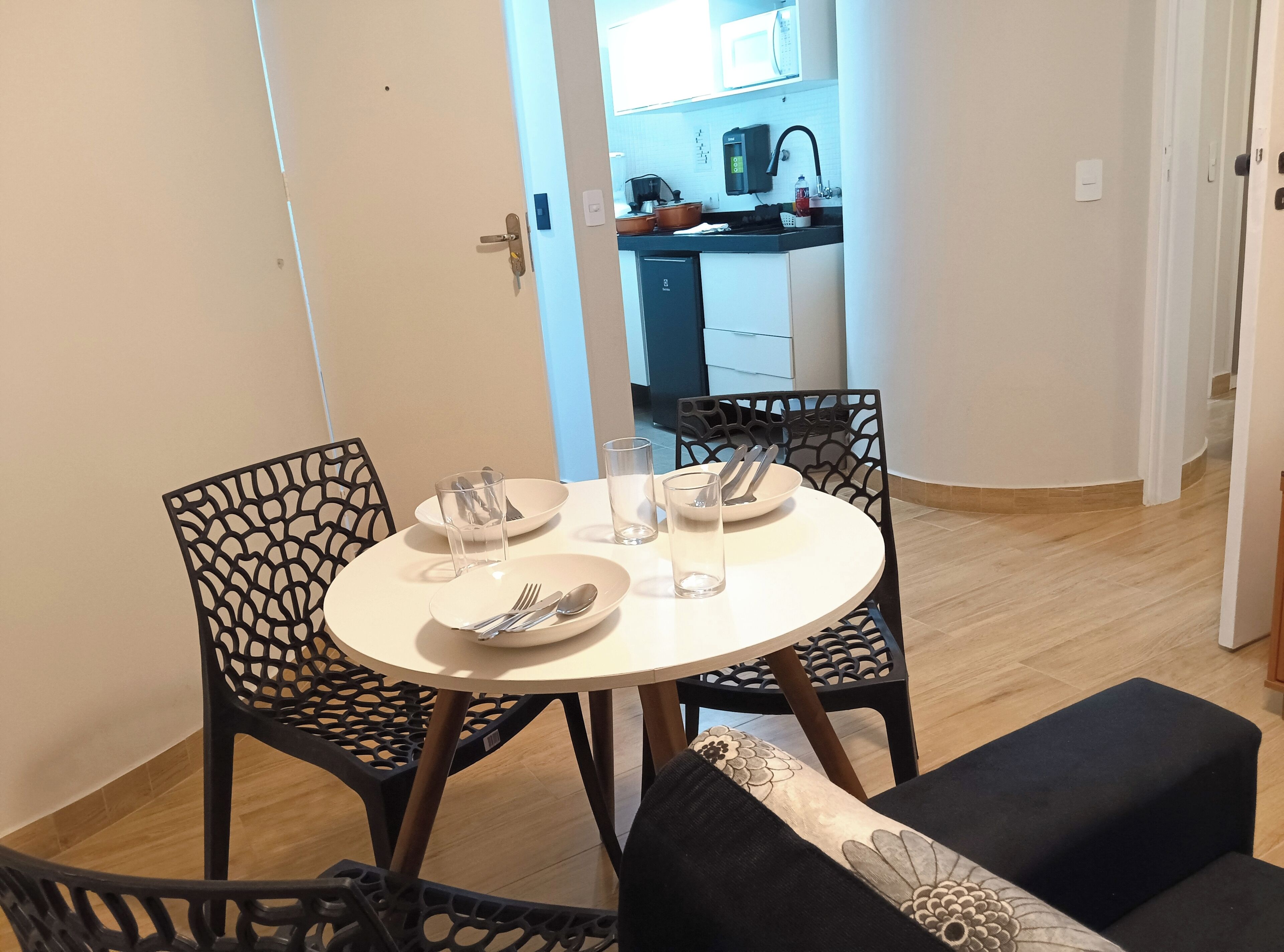 A dining area with a round table, two chairs, and a black ottoman. A kitchen is visible in the background.