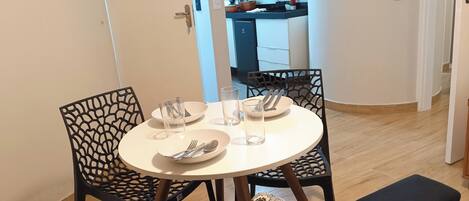 Un coin repas avec une table ronde, deux chaises et un pouf noir. On aperçoit une cuisine au fond.