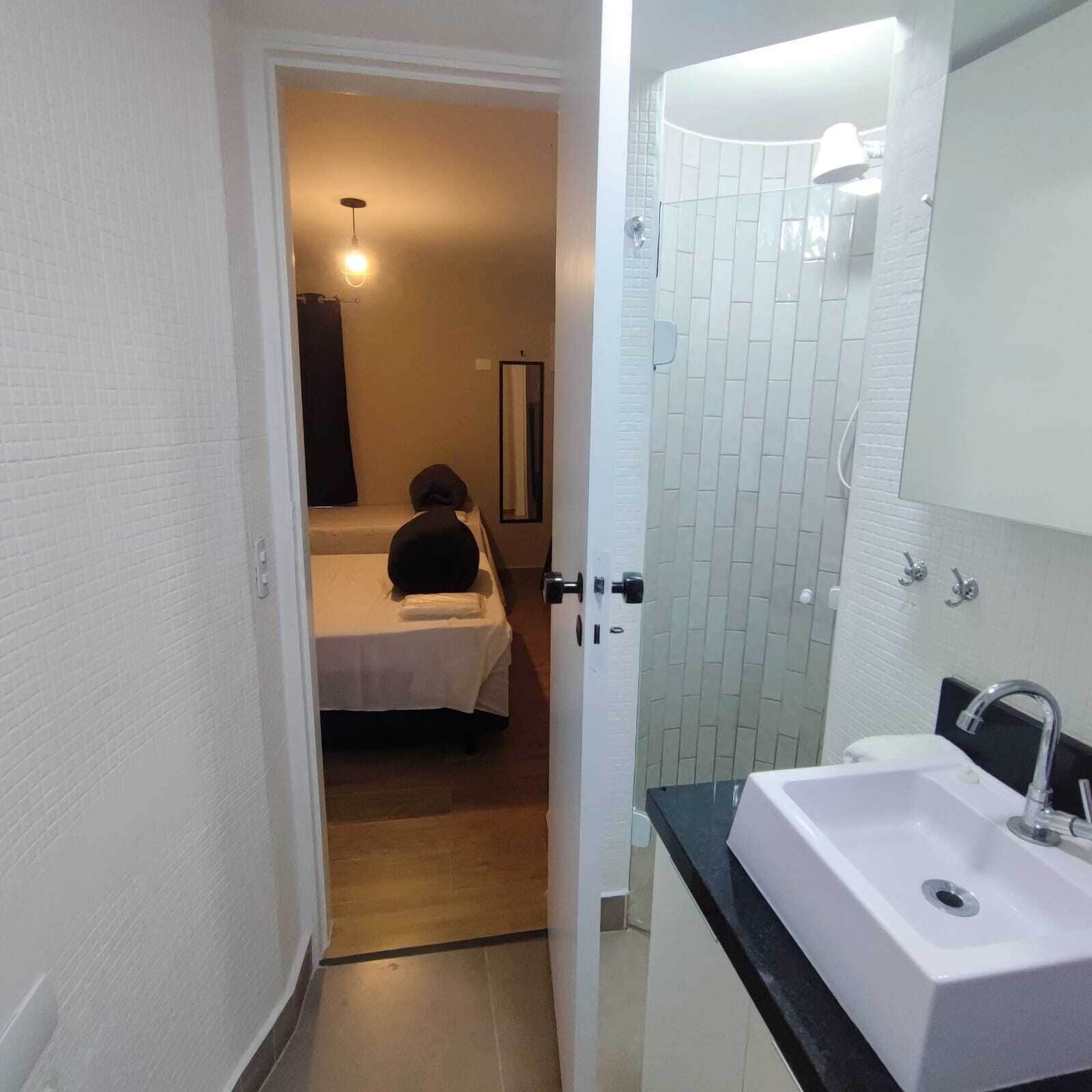 Quarto de hotel com cama, banheiro com pia e espelho, e porta que dá para outro cômodo.