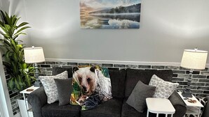 Ein modernes Wohnzimmer mit einem grauen Sofa, einem kleinen weißen Beistelltisch, einem grauen Teppich und einem großen Fenster.