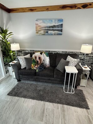 A modern living room with a grey sofa, a small white side table, a grey rug, and a large window. - River Retreat “R&R” Water Front on The St. John’s River (Astor)