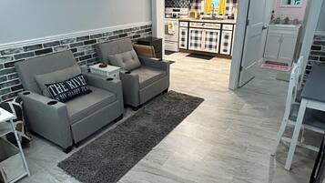 Un salon avec un canapé gris, un tapis et une cuisine en arrière-plan.