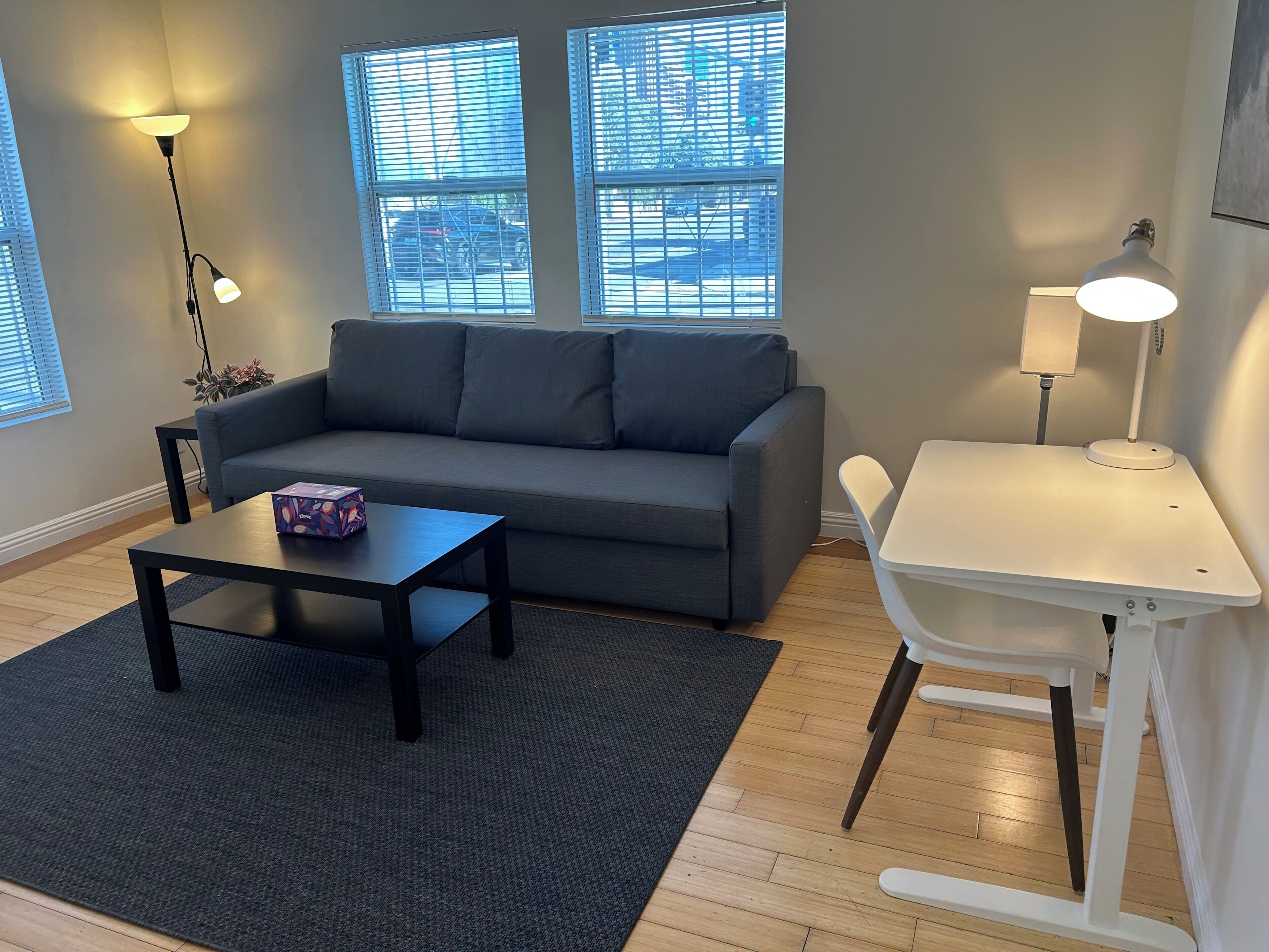 Un salon moderne avec un canapé gris, une petite table noire, un bureau blanc et un lampadaire.