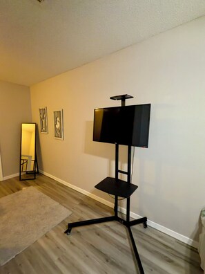 A room with a standing TV on a black stand, a floor lamp, and two framed pictures on the wall. -  2Bed/2Bath Coastal Cove condo, Large pool! Minutes from Gulf Shores sandy coast (Gulf Shores)
