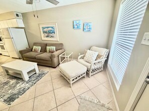 A living room with a sofa, armchair, coffee table, and a small side table. There is a painting on the wall and a window with blinds. -  2Bed/2Bath Coastal Cove condo, Large pool! Minutes from Gulf Shores sandy coast (Gulf Shores)
