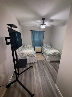 Une chambre avec deux lits, un ventilateur de plafond, un téléviseur fixé sur un support et une petite table.