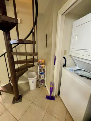 A laundry area with a washing machine, a mop, a trash bin, and a metal shelf with books. -  2Bed/2Bath Coastal Cove condo, Large pool! Minutes from Gulf Shores sandy coast (Gulf Shores)