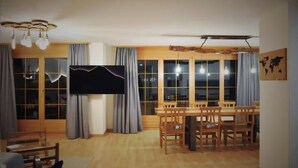 Ein Essbereich mit einem großen Tisch, Stühlen, einem Flachbildfernseher und großen, mit Vorhängen versehenen Fenstern.