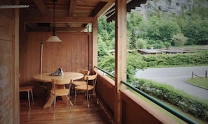 Een houten interieur met een ronde tafel en stoelen, een hanglamp en uitzicht op een groen landschap met een weg en gebouwen.