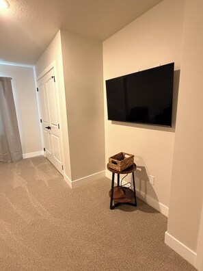 A room with a flat-screen TV mounted on the wall, a small stool with a basket, and a door with a white frame. - 4-bedroom new house in adorable Cedar City with furnished basement (Cedar City)