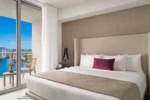 A modern hotel room with a large bed, a view of a cityscape, and a balcony with a chair and a plant.