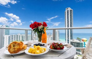 A table with breakfast items, a vase of red roses, and a view of a cityscape and beach.
