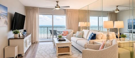A modern living room with a sofa, armchairs, a coffee table, and a TV. There is a balcony with a view of the sea.