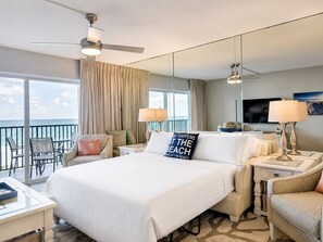 Een hotelkamer met een groot bed, twee nachtkastjes met lampen, een televisie, een balkon met uitzicht op de oceaan en een plafondventilator.
