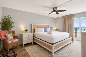 A hotel room with a bed, a nightstand, a chair, a ceiling fan, a window with a view of the beach, and a plant. - BEACHFRONT SUNSETS TO DIE FOR (Madeira Beach)