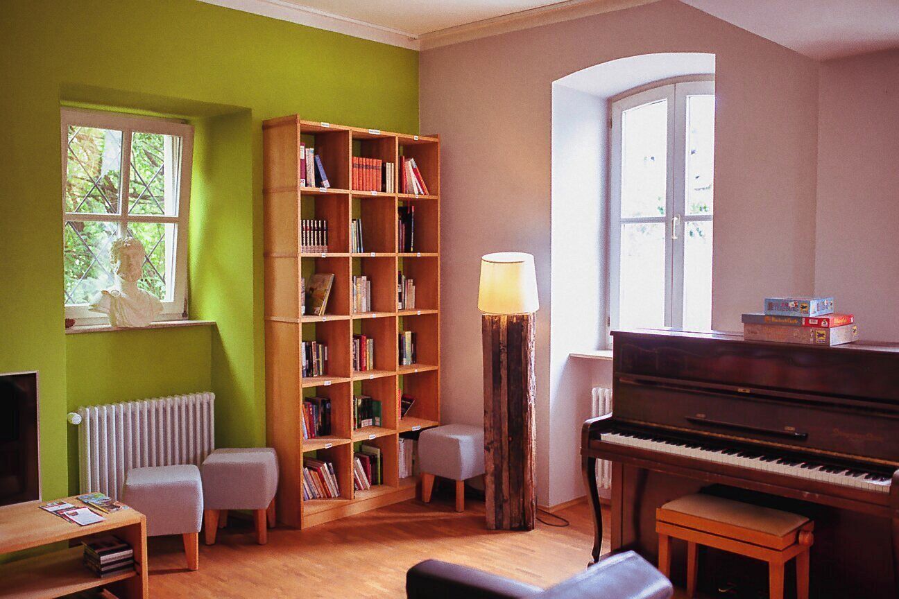 Ein Zimmer mit einem Klavier, einem Bücherregal, einer Lampe und einem Fenster.
