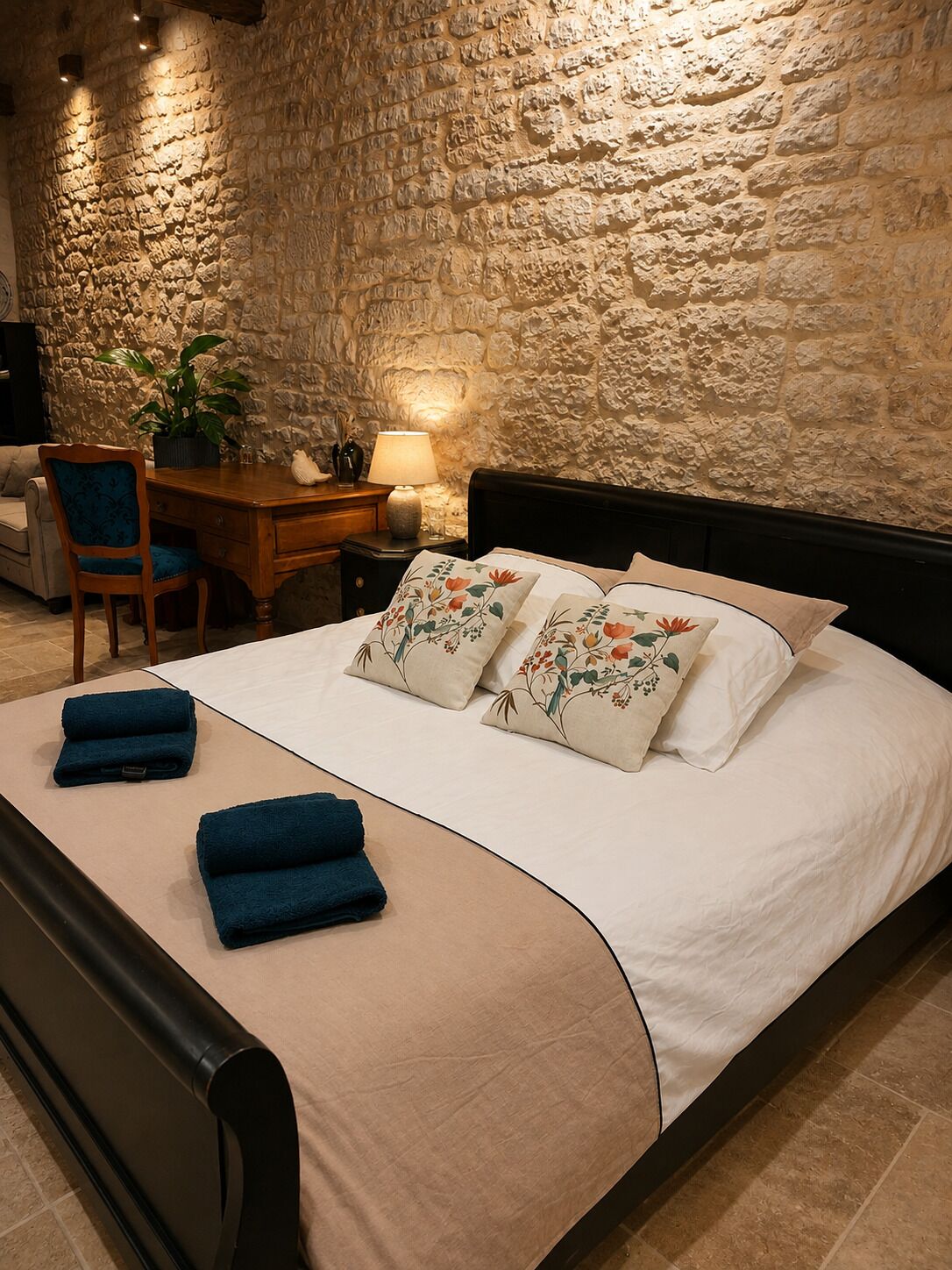 A bedroom with a stone wall, a bed with floral pillows, a desk with a lamp, and a chair.