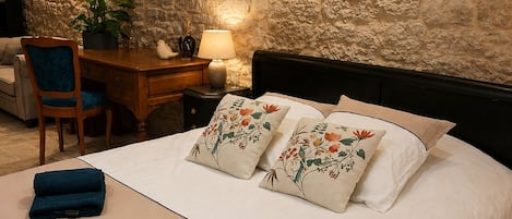 Un dormitorio con pared de piedra, cama con almohadas florales, escritorio con lámpara y una silla.